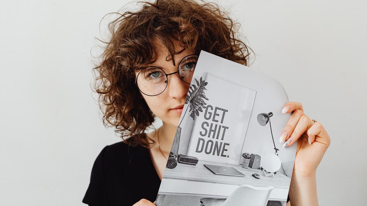 Woman with curly hair and glasses holding a motivational poster saying 'Get Shit Done'